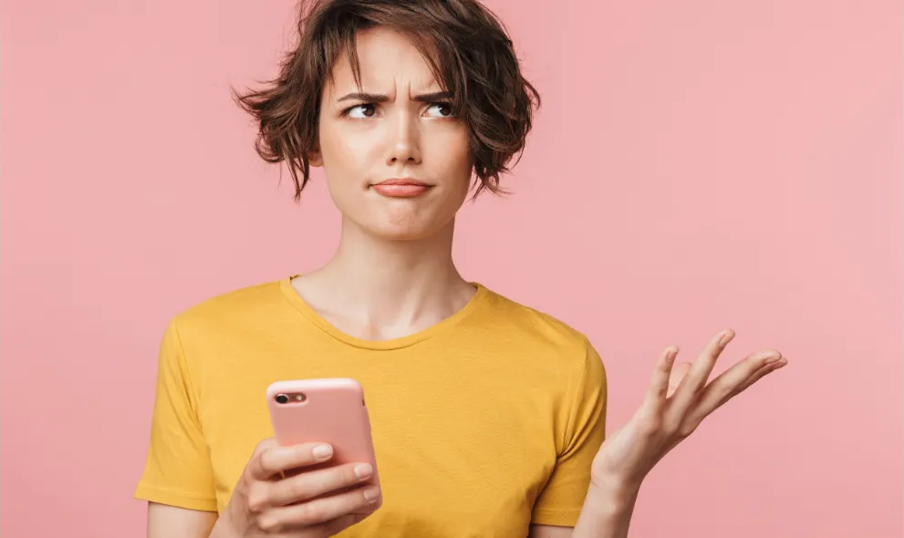 Young confused woman using smartphone on pink background